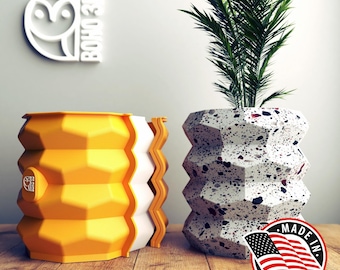 May include: Two modern planters, one yellow and one white with black and red speckles. The white planter has a plant with green leaves. Both planters have a geometric design. The white planter has a sticker that says "Made in USA".
