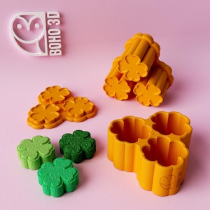May include: An assortment of yellow and green clover-shaped objects, including molds and finished pieces, are arranged on a pink surface. The objects are likely for crafting or baking, with a logo that says "BOHO 3D".