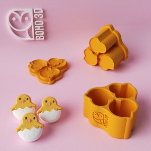 May include: Three yellow chick-shaped silicone molds with a white chick inside each mold. The molds are on a pink surface. There are two other yellow silicone molds in the background, one with three round cavities and the other with three heart-shaped cavities. The molds are labeled "BOHO 3D".