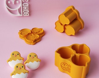 May include: Three yellow chick-shaped silicone molds with a white chick inside each mold. The molds are on a pink surface. There are two other yellow silicone molds in the background, one with three round cavities and the other with three heart-shaped cavities. The molds are labeled "BOHO 3D".