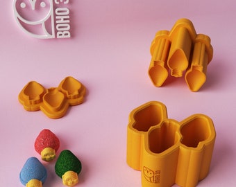 May include: A set of three yellow plastic cookie cutters shaped like Christmas lights. The cutters are on a pink surface. There are three small Christmas light-shaped cookies in red, green, and blue.