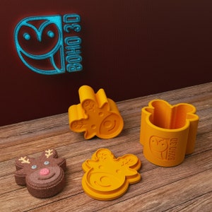 May include: Three orange plastic reindeer cookie cutters, one with a brown reindeer cookie inside. The cookie cutters have a raised design of a reindeer with antlers and a snowflake. The cookie cutter on the right has the text "BOHO 3D" on the side.