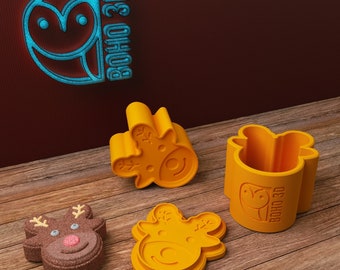 May include: Three orange plastic reindeer cookie cutters, one with a brown reindeer cookie inside. The cookie cutters have a raised design of a reindeer with antlers and a snowflake. The cookie cutter on the right has the text "BOHO 3D" on the side.