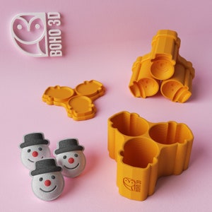 Puede incluir: Tres moldes de plástico amarillo en forma de muñeco de nieve con una cara sonriente. Los moldes están apilados uno encima del otro. Hay tres pequeños caramelos en forma de muñeco de nieve blancos con sombreros negros y narices rojas. También hay un molde de plástico amarillo con tres cavidades en forma de muñeco de nieve. El molde tiene el texto "BOHO 3D" en la parte inferior.