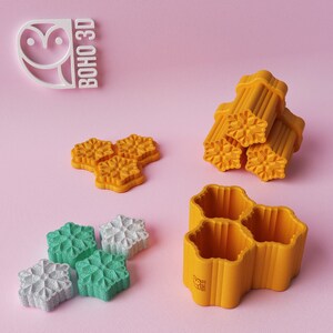 May include: A set of 3D printed cookie cutters in the shape of snowflakes. The cutters are yellow and have a textured surface. The cutters are arranged in a pile and on a pink surface. The image also includes a white owl logo with the text "BOHO 3D".