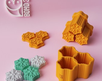 May include: A set of 3D printed cookie cutters in the shape of snowflakes. The cutters are yellow and have a textured surface. The cutters are arranged in a pile and on a pink surface. The image also includes a white owl logo with the text "BOHO 3D".