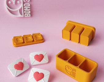 May include: Three small white envelopes with red hearts on them, and three yellow plastic containers with the logo "BOHO 3D" on them. The containers are designed to hold the envelopes.