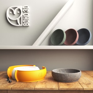 May include: The image showcases a variety of handcrafted bowls and soap dishes. There are three round soap dishes in green, pink, and blue. Two orange and white bowls are also visible, along with a gray stone bowl. The Boho 3D logo is displayed on the wall.