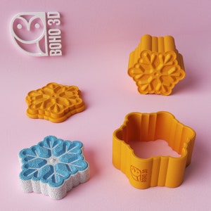 May include: A collection of 3D-printed snowflake-shaped cookie cutters in yellow and blue, with a white owl logo that reads "BOHO 3D" on a pink background. The cutters have detailed snowflake designs, perfect for baking seasonal treats.