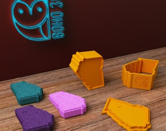 May include: Five different colored cookie cutters shaped like the state of Georgia. The cookie cutters are on a wooden surface. The cookie cutters are made of plastic and have a 3D design. The cookie cutters are all different colors, including teal, purple, pink, orange, and yellow.