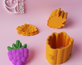 May include: Four 3D printed molds for making raspberry-shaped treats. The molds are yellow and have a textured surface. One mold is a single raspberry shape, while the other three are stacked together to form a larger raspberry shape. The molds are on a pink surface. The text "BOHO 3D" is visible on one of the molds.