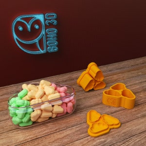 May include: Three orange plastic heart-shaped cookie cutters, one stacked on top of the other, and two separate cutters. A glass container filled with pastel-colored heart-shaped candies. The container and cutters are on a wooden surface. The text "BOHO 3D" is visible in the background.