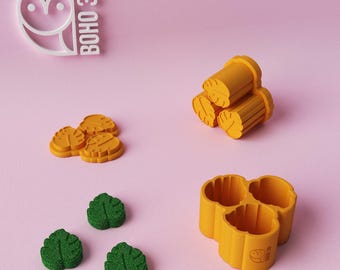 May include: A collection of 3D-printed cookie cutters in yellow and green, including leaf and log shapes, and a three-sectioned mold. The Boho 3D logo is visible in the upper left corner. The objects are arranged on a pink background.