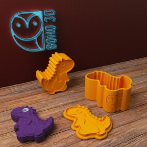 May include: Four dinosaur-shaped cookie cutters, two yellow and two purple. The yellow cutters are in the shape of a standing dinosaur and a dinosaur lying down. The purple cutters are in the shape of a standing dinosaur. The cutters are on a wooden surface. The text "BOHO 3D" is visible on one of the cutters.