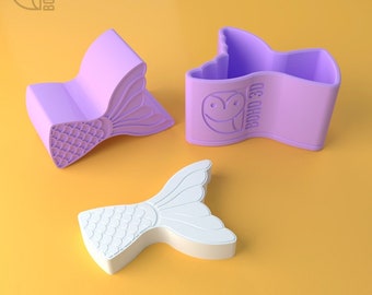 May include: Three mermaid tail molds, two are purple and one is white. The molds are made of silicone and have a detailed mermaid tail design. The molds are for making soap, candles, or other crafts.