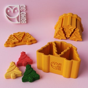 May include: A set of 3D printed yellow Christmas tree cookie cutters. The cutters are in the shape of a Christmas tree and have a star on top. The cutters are arranged in a stack, with the largest cutter on the bottom and the smallest cutter on top. There are also three small Christmas tree-shaped cookies in red, green, and yellow.
