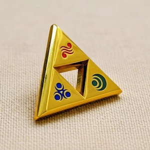 Puede incluir: Pin triangular dorado con un centro recortado y tres triángulos más pequeños, cada uno con un diseño diferente en rojo, azul y verde. El pin es probablemente un objeto de colección o un accesorio.