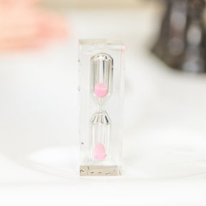 Könnte beinhalten: Eine klare Sanduhr mit rosa Sand, die auf einer weißen Oberfläche steht.