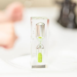 Könnte beinhalten: Eine klare Sanduhr mit silbernem Rahmen und grünem Sand. Die Sanduhr steht auf einer weißen Oberfläche.