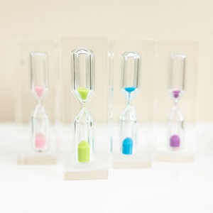 Könnte beinhalten: Vier transparente Glas-Sanduhren mit farbigem Sand in Rosa, Grün, Blau und Lila. Jede Sanduhr ist in einem transparenten rechteckigen Block eingeschlossen. Der Sand befindet sich am Boden.