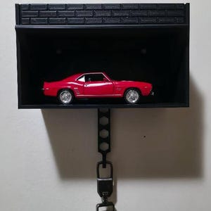 May include: A red toy car is displayed inside a black wall-mounted shelf. The shelf has a textured top and is connected to a key fob by a black chain. The key fob is black with silver buttons and a key symbol.