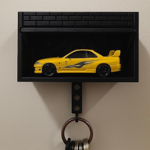 May include: A black key holder shaped like a garage with a yellow toy car inside. The car has a black racing stripe and the number "7" on the side. The key holder has a metal hook for hanging keys.