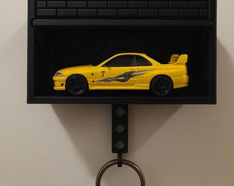 May include: A black key holder shaped like a garage with a yellow toy car inside. The car has a black racing stripe and the number "7" on the side. The key holder has a metal hook for hanging keys.