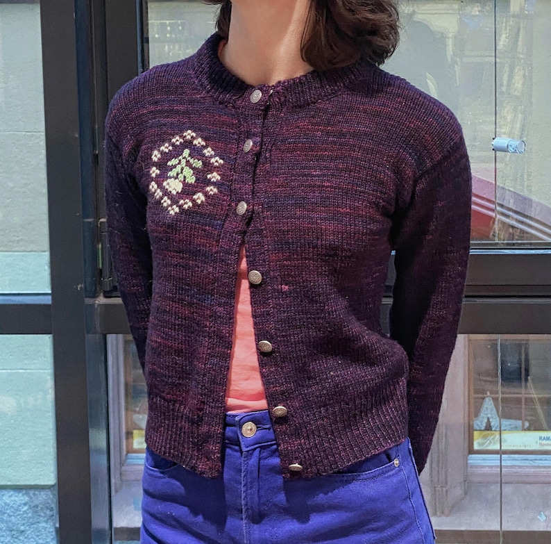 Peut inclure: Un cardigan en tricot violet et noir avec un motif brod&eacute; floral sur le c&ocirc;t&eacute; gauche. Le cardigan a une fermeture boutonn&eacute;e sur le devant et des manches longues.