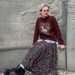 May include: A woman wearing a maroon sweater with a white embroidered deer design, a black floral skirt, and black boots. She is sitting on a stone bench in front of a stone monument with carved text.