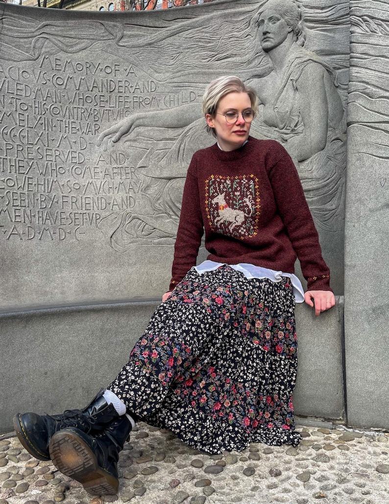 May include: A woman wearing a maroon sweater with a white embroidered deer design, a black floral skirt, and black boots. She is sitting on a stone bench in front of a stone monument with carved text.