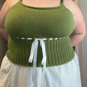K&ouml;nnte beinhalten: Ein gr&uuml;nes Stricktop mit einer wei&szlig;en Schleife an der Taille. Das Top wird &uuml;ber einem wei&szlig;en Spitzenrock getragen.