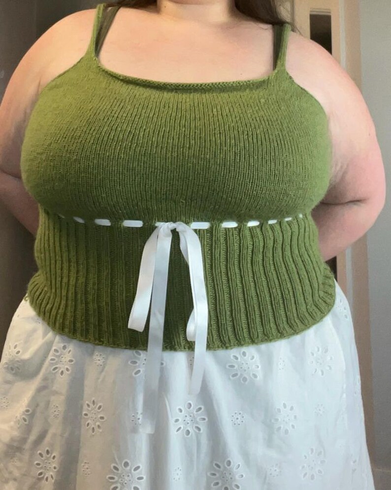 K&ouml;nnte beinhalten: Ein gr&uuml;nes Stricktop mit einer wei&szlig;en Schleife an der Taille. Das Top wird &uuml;ber einem wei&szlig;en Spitzenrock getragen.