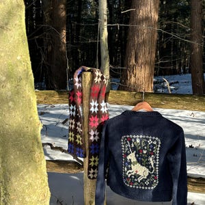 May include: A navy blue knitted cardigan with a white unicorn and floral design on the back. A brown, pink, blue, and yellow knitted scarf is hanging on a wooden post in the background.