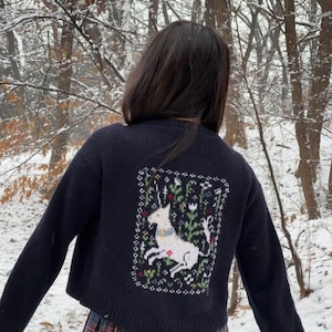 Peut inclure: Un cardigan bleu marine court avec une licorne blanche brod&eacute;e au dos. Le cardigan est en tricot torsad&eacute; et se ferme par des boutons.