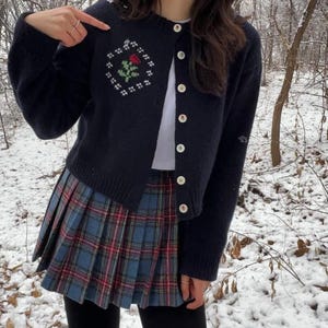 Peut inclure: Un cardigan bleu marine avec une rose blanche brod&eacute;e sur le devant. Le cardigan a une fermeture boutonn&eacute;e et est port&eacute; sur un t-shirt blanc et une jupe &agrave; carreaux bleus et rouges.