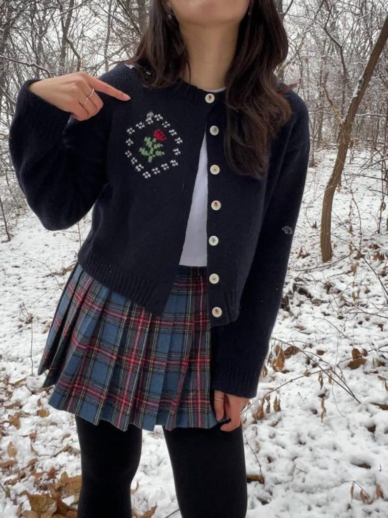 Peut inclure: Un cardigan bleu marine avec une rose blanche brod&eacute;e sur le devant. Le cardigan a une fermeture boutonn&eacute;e et est port&eacute; sur un t-shirt blanc et une jupe &agrave; carreaux bleus et rouges.