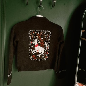 Peut inclure: Un cardigan marron avec une licorne blanche brod&eacute;e au dos. La licorne est entour&eacute;e de fleurs et de feuilles dans des tons de rouge, vert et jaune.