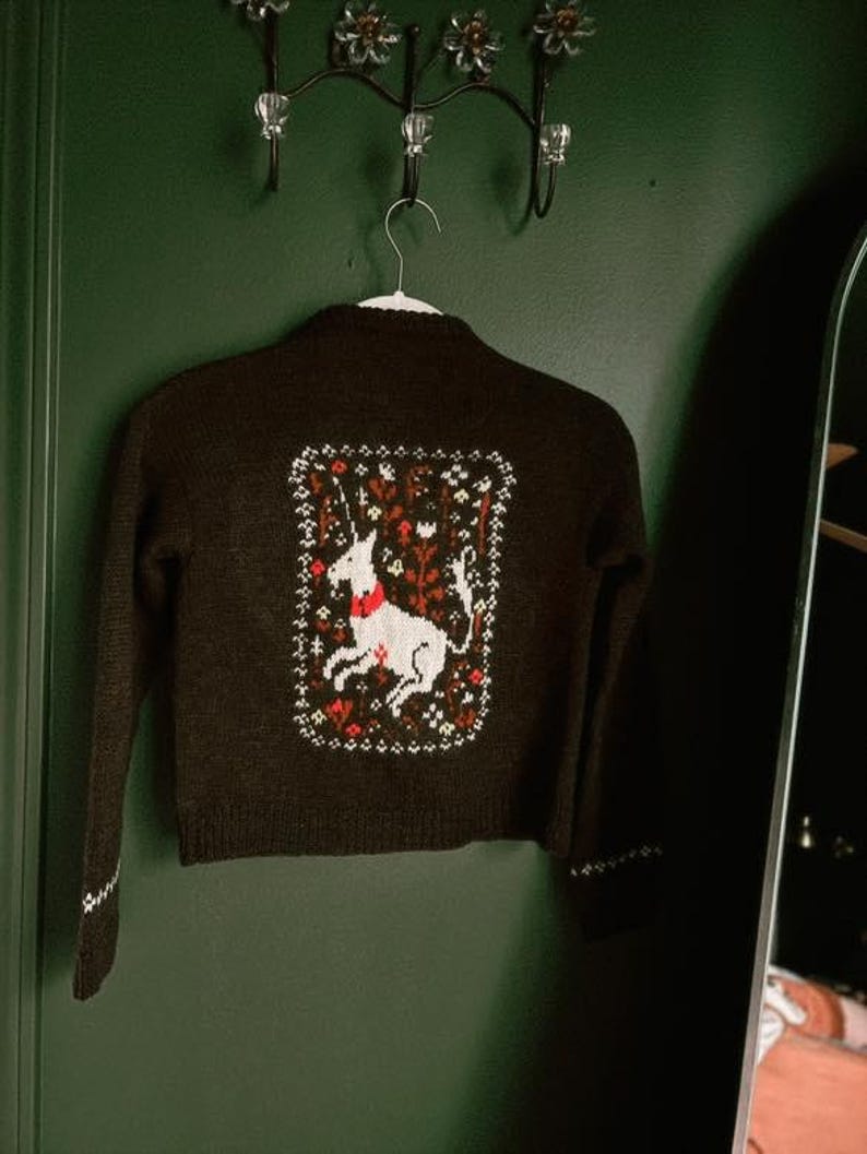 Peut inclure: Un cardigan marron avec une licorne blanche brod&eacute;e au dos. La licorne est entour&eacute;e de fleurs et de feuilles dans des tons de rouge, vert et jaune.