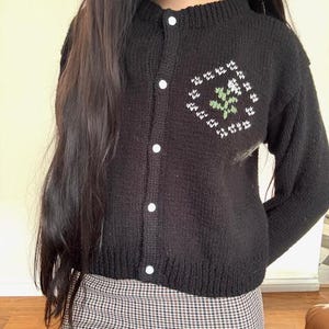 Peut inclure: Un cardigan en tricot noir avec des boutons blancs et un motif de fleur brod&eacute; en vert et blanc.