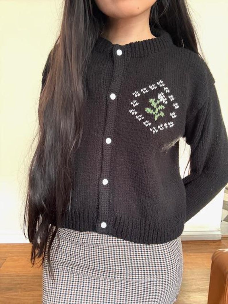 Peut inclure: Un cardigan en tricot noir avec des boutons blancs et un motif de fleur brod&eacute; en vert et blanc.
