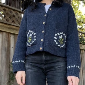 Peut inclure: Un cardigan bleu fonc&eacute; avec des broderies florales blanches et vertes sur les poches. Le cardigan a une fermeture boutonn&eacute;e &agrave; l'avant et des manches longues.