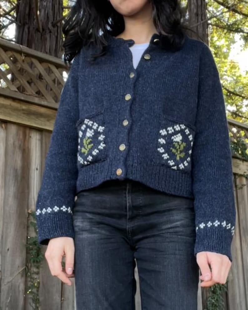 Peut inclure: Un cardigan bleu fonc&eacute; avec des broderies florales blanches et vertes sur les poches. Le cardigan a une fermeture boutonn&eacute;e &agrave; l'avant et des manches longues.