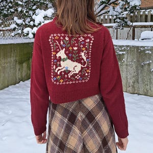 Peut inclure: Un cardigan rouge court avec une licorne blanche brod&eacute;e au dos. Le cardigan est en tricot torsad&eacute; et est port&eacute; sur une jupe &agrave; carreaux.