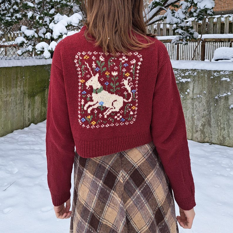 Peut inclure: Un cardigan rouge court avec une licorne blanche brod&eacute;e au dos. Le cardigan est en tricot torsad&eacute; et est port&eacute; sur une jupe &agrave; carreaux.