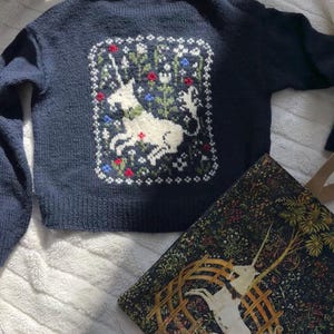May include: A navy blue knitted sweater with a white unicorn embroidered on the back. The unicorn is surrounded by green leaves and red, white, and blue flowers.