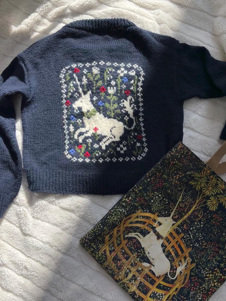 Peut inclure: Un pull en tricot bleu marine avec une licorne blanche brod&eacute;e au dos. La licorne est entour&eacute;e de feuilles vertes et de fleurs rouges, blanches et bleues.