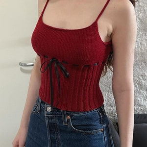 K&ouml;nnte beinhalten: Ein rotes Stricktop mit einem schwarzen Schleifenband vorne. Das Top hat eine figurbetonte Silhouette und eine gerippte Textur.