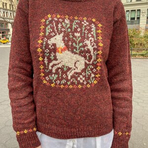 May include: A maroon sweater with a white unicorn and floral design in the center. The sweater has a crew neck and long sleeves.