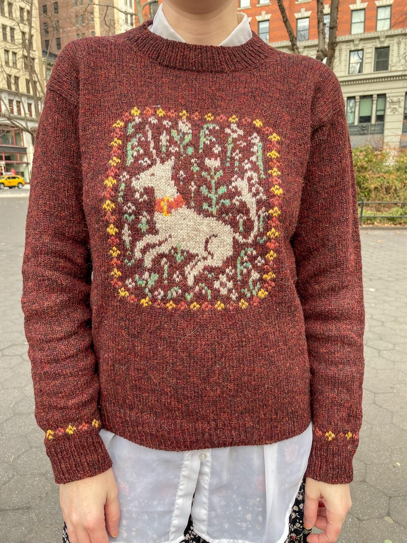 May include: A maroon sweater with a white unicorn and floral design in the center. The sweater has a crew neck and long sleeves.