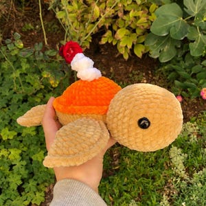 Könnte beinhalten: Ein handgefertigtes Schildkrötenspielzeug mit einem strukturierten, beigefarbenen Körper, einem orangefarbenen Panzer und einem roten und weißen Dekorationselement. Die Schildkröte hat ein schwarzes Knopfauge und wird in einer Hand gehalten.